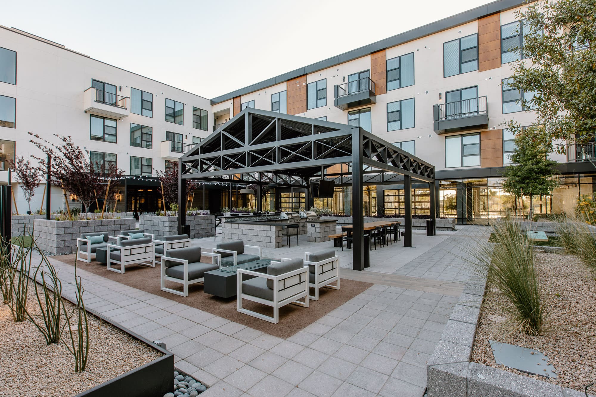 Modern apartment complex courtyard featuring neatly arranged patio furniture under a large pergola, surrounded by well-maintained garden areas.