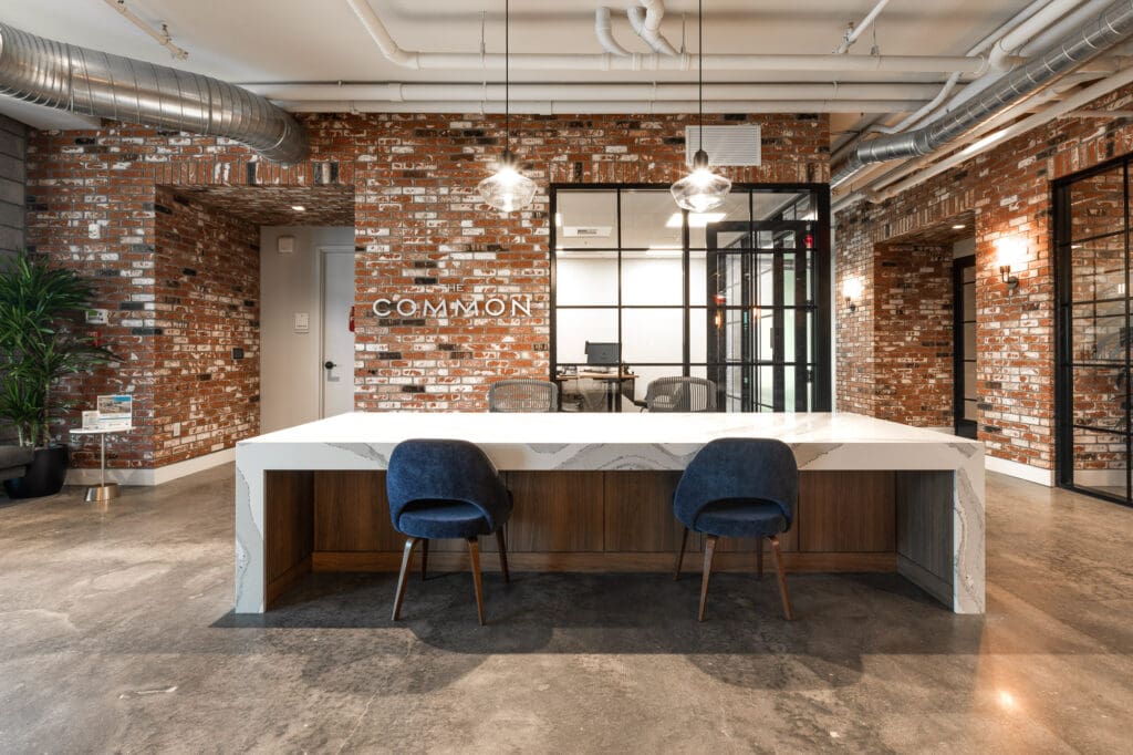 Modern office reception area featuring a white desk with two blue chairs, exposed brick walls, and industrial-style ceiling.