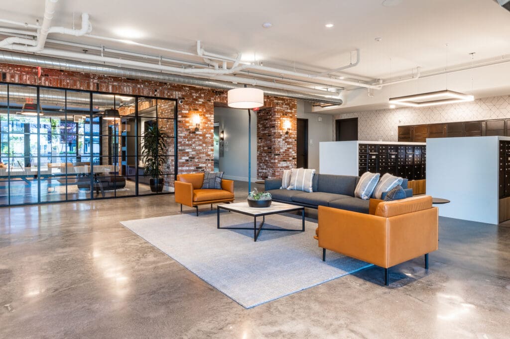 Modern office lobby with exposed brick walls, gray and orange sofas, a central coffee table, and polished concrete floors, illuminated by pendant and recessed lighting.