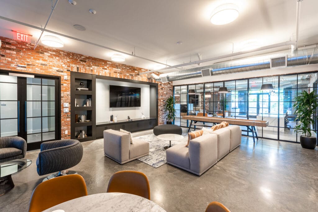 Modern office lounge area with sofas, exposed brick walls, and a large screen tv, illuminated by natural light from large windows.