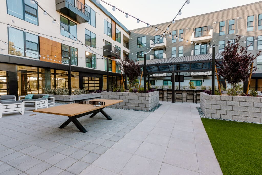 A modern apartment courtyard with benches, a ping pong table, and string lights, surrounded by lush landscaping and stylish residential buildings.