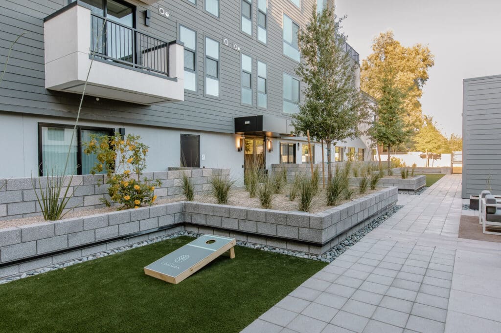 Modern apartment building with a landscaped courtyard featuring raised garden beds, a cornhole game on synthetic grass, and seating areas.