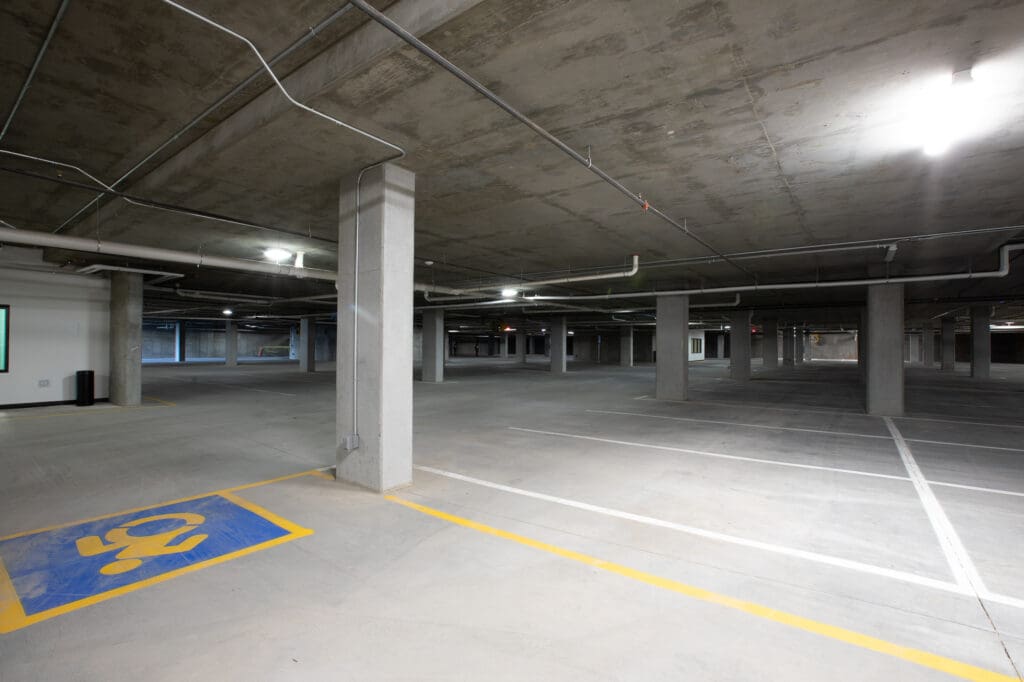 Empty underground parking garage with painted lines, some pillars, and overhead lighting. handicap parking space visible.