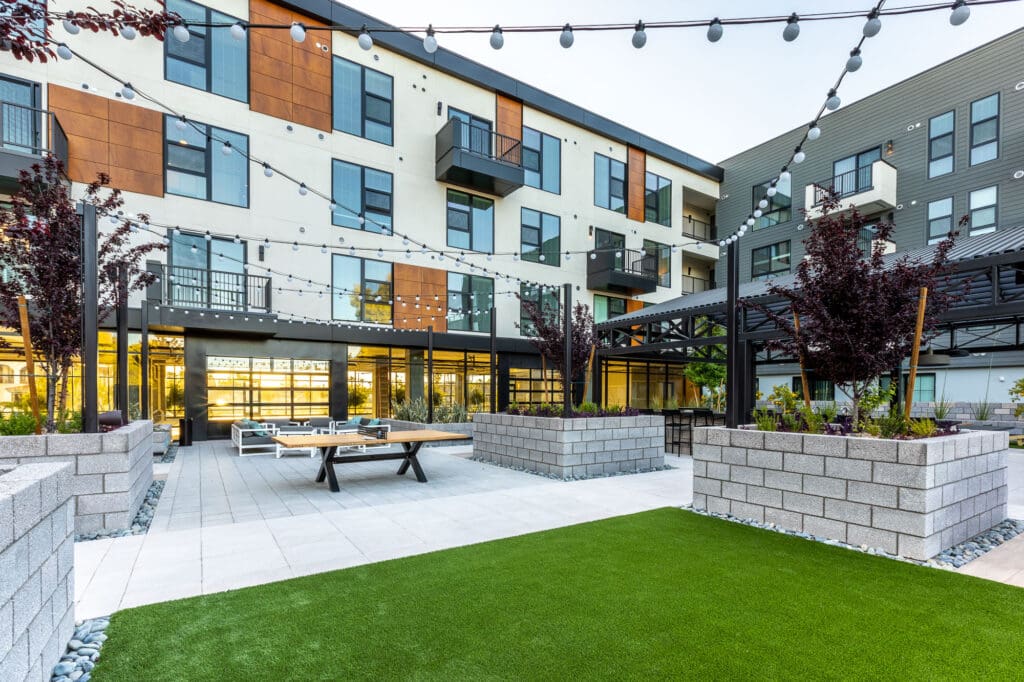 Modern apartment building courtyard with green artificial grass, string lights, picnic tables, and surrounding residential units with large glass windows.
