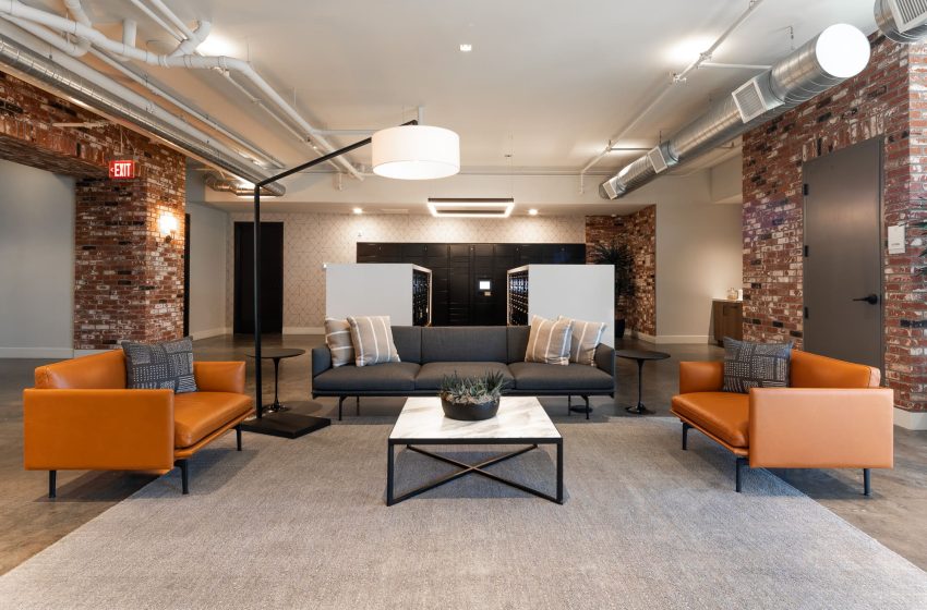 Modern office lobby with exposed brick walls, two orange armchairs, a gray sofa, and a central coffee table on a large area rug, under white ceiling lights.