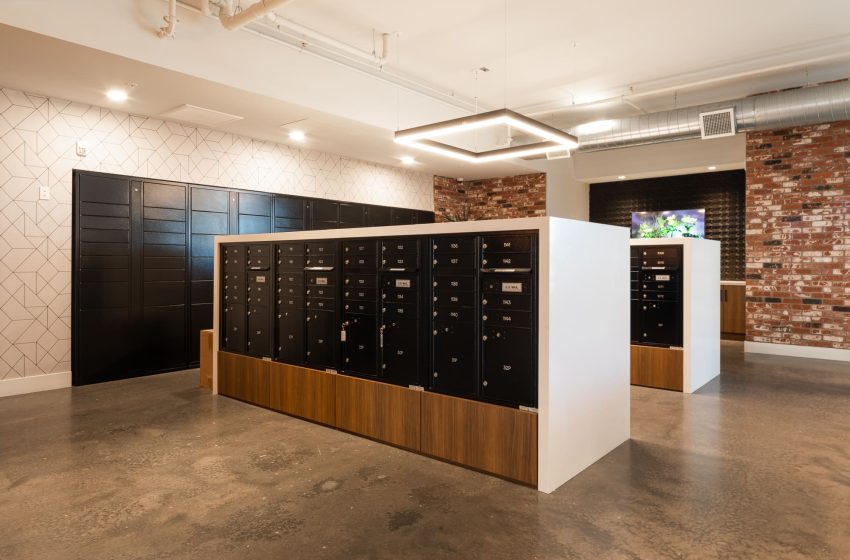 Modern office lobby featuring a central block of black mailboxes with wood paneling, surrounded by exposed brick and patterned tile walls, under bright ceiling lights.