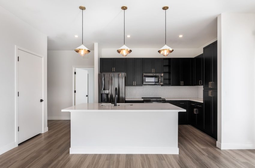 Modern kitchen with white countertops, black cabinets, and stainless steel appliances, featuring a central island and pendant lights.