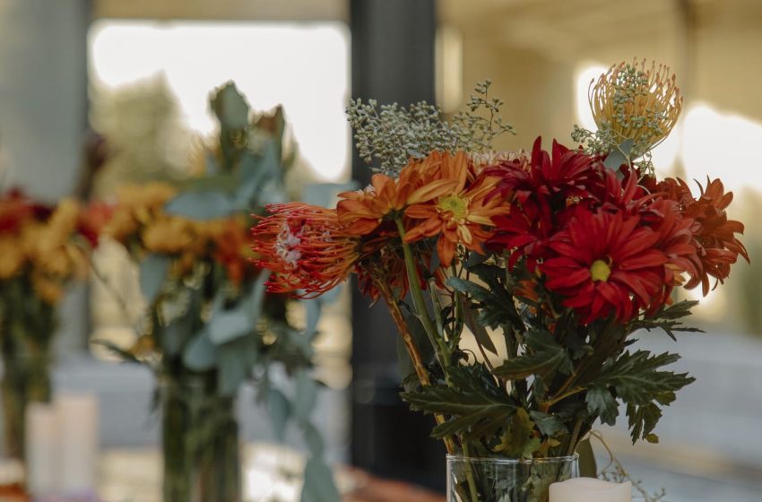 A row of autumnal flower arrangements with red and orange blooms, accompanied by candles and small pumpkins, set on a woven table runner.