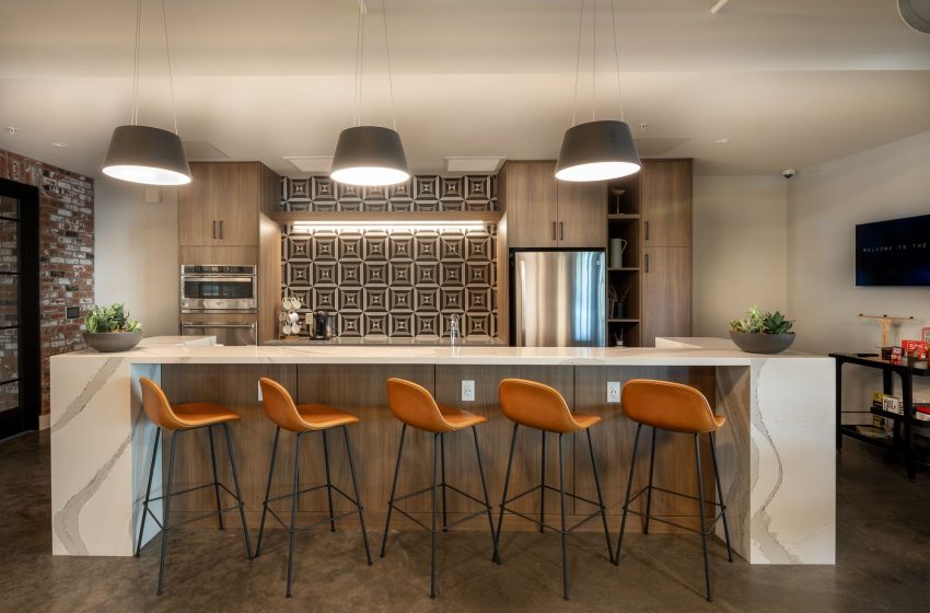 Modern kitchen with exposed brick, a large island with orange stools, pendant lights, stainless steel appliances, and wooden cabinetry.