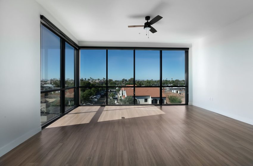 Empty modern living room with large windows showing a residential area and clear sky, wooden flooring, and a ceiling fan.