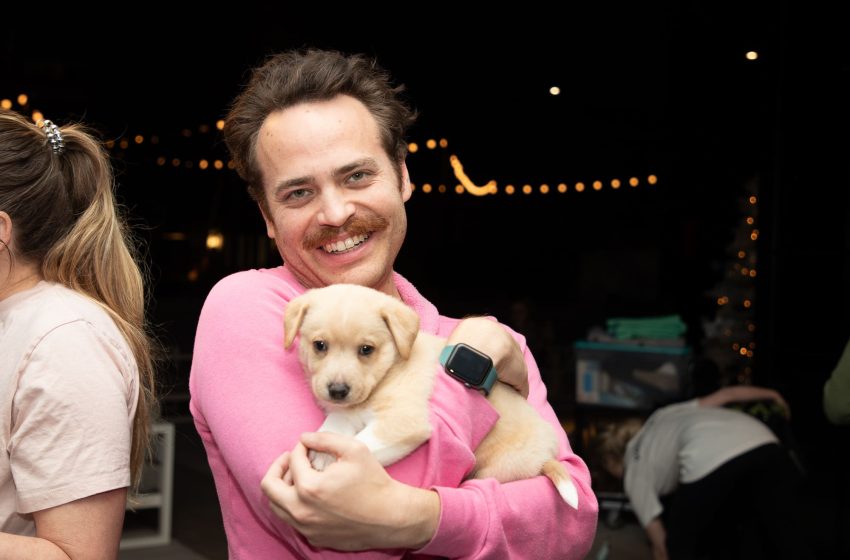 A smiling man in a pink shirt holding a small golden puppy at an outdoor event with string lights in the background.