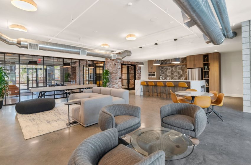 Modern office space featuring a lounge area with sofas, a communal worktable, brick walls, ductwork on ceiling, and large windows.