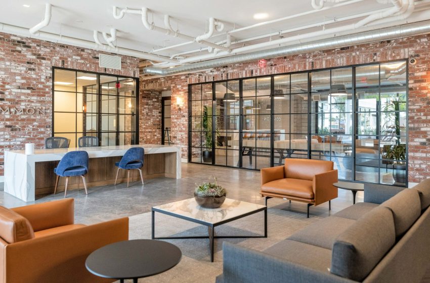 Modern office lobby with exposed brick walls, glass partition doors, leather sofas, a reception desk, and stylish hanging lights.