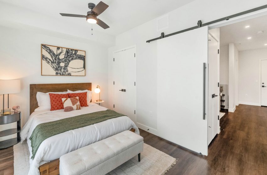 Modern bedroom featuring a queen-sized bed with green and orange bedding, white sliding doors, hardwood floors, and abstract art above the bed.