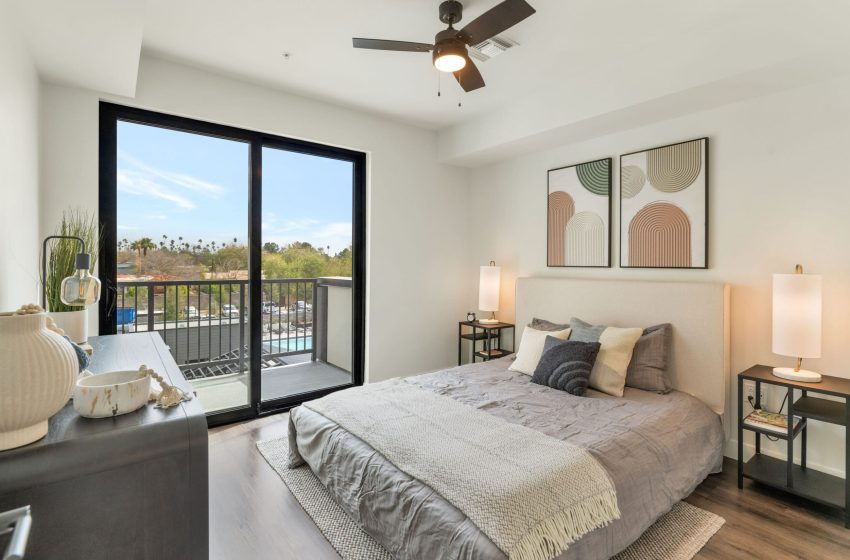 Modern bedroom with a large bed, two nightstands with lamps, artworks on the wall, and a ceiling fan, featuring a balcony that overlooks a palm tree landscape.