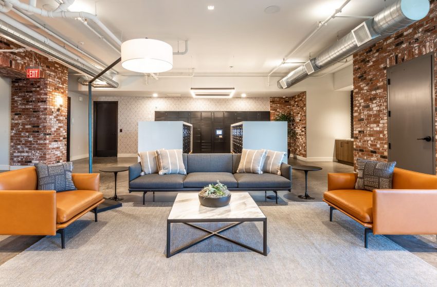 Modern office lobby with exposed brick walls, a blue sofa, two orange leather chairs, a white coffee table, and industrial-style ceiling.